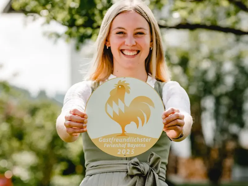 Gastgeberin mit Goldener Gockel Plakette - © Kimpelehof Eine Gastgeberin mit ihrer Plakette der Auszeichnung Goldener Gockel - Gastfreundlichster Ferienhof Bayerns im Dirndl im Garten ihres Urlaubsbauernhofes.